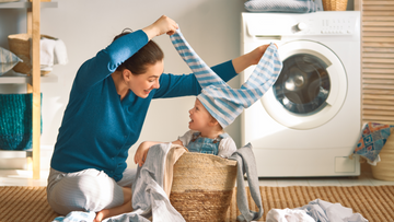 Moeder en kind spelen samen met wasgoed naast een wasmachine – een vrolijk moment tijdens natuurlijk en veilig wassen thuis.